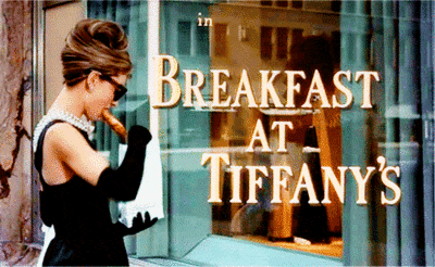 Audrey Hepburn as Holly stands outside the Tiffany's shop window with coffee and a pretzel.